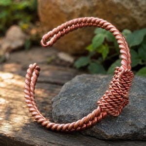 Puede incluir: Una pulsera tipo brazalete de cobre con un diseño de alambre trenzado. La pulsera está construida con múltiples hilos de alambre de cobre metálico que están enrollados y envueltos juntos para formar una banda circular abierta y continua.