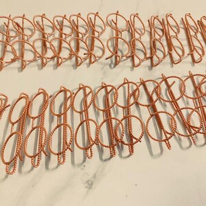 May include: Rows of copper-colored hair barrettes. Each barrette features a twisted wire design with a circular top and a vertical stem. The barrettes are arranged in neat rows, showcasing their intricate details and metallic sheen.