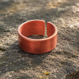 Puede incluir: Un anillo de cobre ancho y abierto con un acabado cepillado y texturizado. La banda presenta un diseño plano y sencillo con una abertura distintiva en la parte superior, mostrando el color marrón rojizo metálico y el aspecto natural.