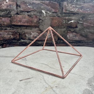 Op de afbeelding: Koperkleurige piramide met een vierkante basis, gemaakt van gedraaide draad. De getextureerde piramide staat op een betonnen oppervlak, met een bakstenen muur op de achtergrond. Afmetingen: ongeveer 10 cm hoog.