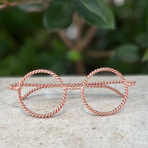 May include: A pair of decorative eyeglasses crafted from a single continuous piece of twisted copper wire. The frame features two circular lenses outlined by the rope-like texture of the metal, connected by a straight bridge and side arms.