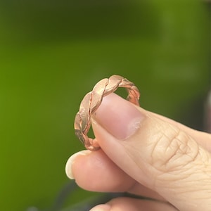 Könnte beinhalten: Ein kupferfarbener Ring mit einem durchgehenden, gedrehten oder geflochtenen Metalldesign. Die Oberfläche des Bandes weist eine texturierte, matte Optik mit sichtbaren Graten auf, die dem ineinandergreifenden Muster um den gesamten Ring folgen.