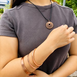 Puede incluir: Un colgante de color cobre con tres círculos concéntricos trenzados en un collar de cordón negro, junto con tres brazaletes de color cobre en la muñeca, que incluyen una banda ancha lisa y dos brazaletes más finos con diseño de cuerda.