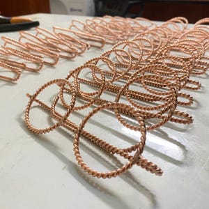 May include: Close-up of several copper-colored metal wire structures. The wire is twisted into a rope-like pattern, forming circular and oval shapes. The objects are arranged on a light-colored surface, suggesting a craft or DIY project.