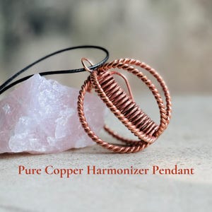 Op de afbeelding: Een hanger met een bolvormige kooi van gedraaid koperdraad met een centrale spoel, hangend aan een zwart koord naast een ruw, lichtroze kristal. De tekst Pure Copper Harmonizer Pendant staat onderaan de afbeelding gedrukt.