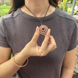 May include: A copper-colored pendant featuring a spiral design made of twisted wire, suspended on a thin black cord. The pendant is accompanied by a matching twisted wire bracelet, both showcasing a metallic finish and a consistent rope-like texture.