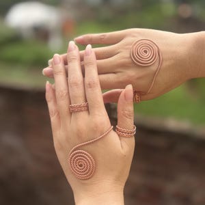 May include: A set of copper-coloured wire jewellery consisting of two rings with textured bands and two hand ornaments featuring a large spiral design that rests on the back of the hand, connected to a ring that encircles the finger with a twisted rope pattern.