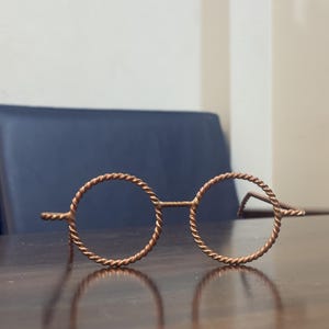 May include: A pair of round, copper-colored wireframe glasses with a twisted rope design. The glasses are resting on a dark wooden surface, with a dark blue chair in the background.