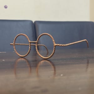May include: A pair of round-framed eyeglasses made from twisted copper wire. The frames and temples have a textured, braided appearance. The glasses are resting on a dark wooden surface, with a blurred background.