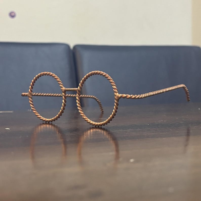 May include: A pair of round-framed eyeglasses made from twisted copper wire. The frames and temples have a textured, braided appearance. The glasses are resting on a dark wooden surface, with a blurred background.