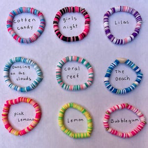 May include: Nine colorful polymer clay bracelets with various color schemes and names like "Cotton Candy," "Girls Night," and "Lilac." Each bracelet is made of small, flat beads. The bracelets are arranged on a white surface.