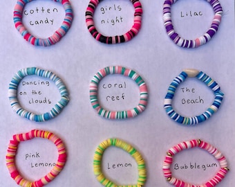 Op de afbeelding: Negen kleurrijke polymeerklei armbanden met namen als "Cotton Candy", "Girls Night" en "Lilac". Elke armband is gemaakt van kleine, platte kralen. De armbanden zijn gerangschikt op een wit oppervlak. Afmetingen niet beschikbaar.