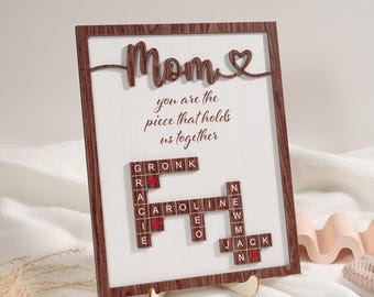 May include: A wooden frame with a white background and a brown wood grain border. The frame has a scrabble tile design with the words "Mom you are the piece that holds us together" and a heart symbol. The tiles are arranged in a crossword puzzle pattern.