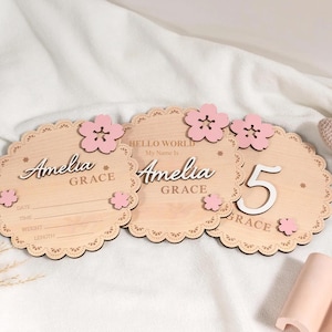 May include: Three wooden plaques with scalloped edges and pink flower accents. The plaques have the name "Amelia Grace" and the text "Hello World My Name Is". One plaque displays the number "5". The plaques are light brown.