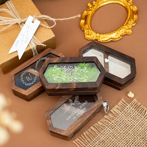 May include: Wooden hexagonal ring boxes with clear lids. Some boxes have names and dates etched on the glass. One box contains green moss. A gift box with a tag that says "Especially for you" is in the background.