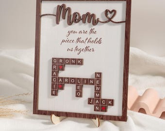 May include: A wooden framed sign with the word "Mom" in cursive with a heart. The sign has a scrabble tile crossword puzzle with the words "GRONK", "CAROLINE", "NEW", "EWM", and "JACK". The sign reads "You are the piece that holds us together".