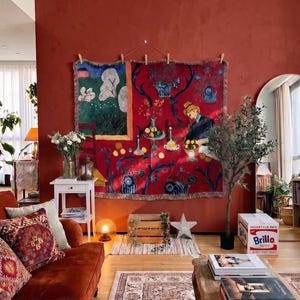 May include: A large rectangular woven wall tapestry featuring a vibrant red background with a painted scene of a person, fruit, and a window view. The tapestry is hung with wooden clips against a deep terracotta wall above a wooden crate.