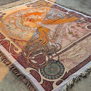 May include: A woven throw blanket featuring an Art Nouveau-style illustration of a woman with long flowing hair. The blanket has a detailed design with warm colors like orange, red, and beige, and a fringed edge.