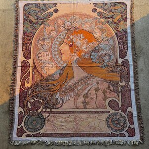 May include: Woven tapestry throw blanket featuring an Art Nouveau style design. The artwork depicts a woman's profile with flowing orange hair, surrounded by an ornate circular frame. The color palette includes shades of peach, burgundy, and teal, with a fringed edge.