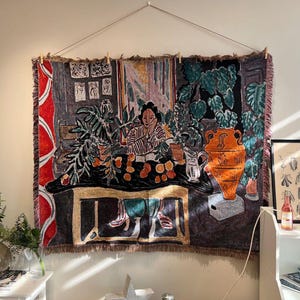 May include: A woven tapestry featuring a woman seated at a table with plants and an orange vase. The artwork has a dark color palette with red, green, and orange accents. The tapestry is hung with wooden clothespins.