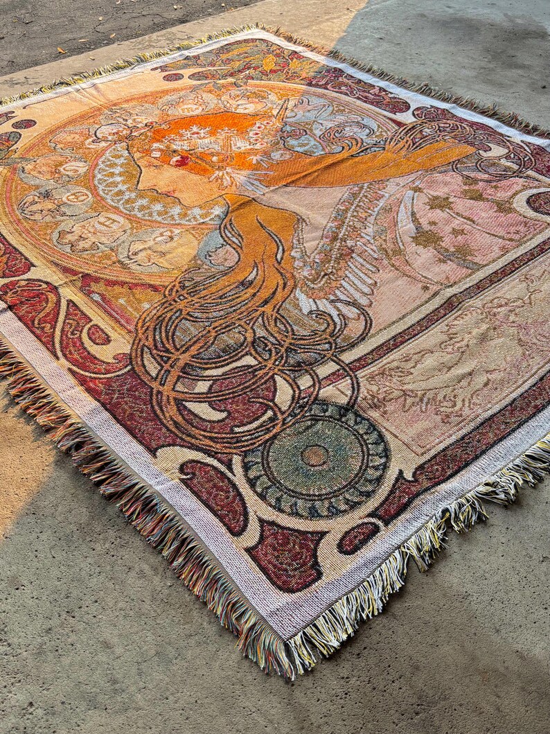 May include: A woven throw blanket featuring an Art Nouveau-style illustration of a woman with long flowing hair. The blanket has a detailed design with warm colors like orange, red, and beige, and a fringed edge.
