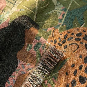 May include: A woven throw blanket with a jungle-themed design. The blanket features a leopard print, black animal silhouette, and green and pink foliage. The fringed edge adds a decorative touch to this cozy home decor item.