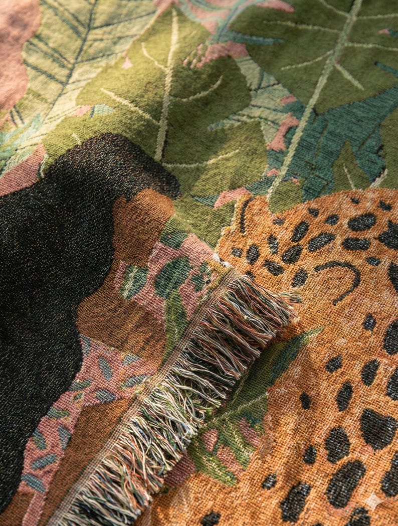 May include: A woven throw blanket with a jungle-themed design. The blanket features a leopard print, black animal silhouette, and green and pink foliage. The fringed edge adds a decorative touch to this cozy home decor item.