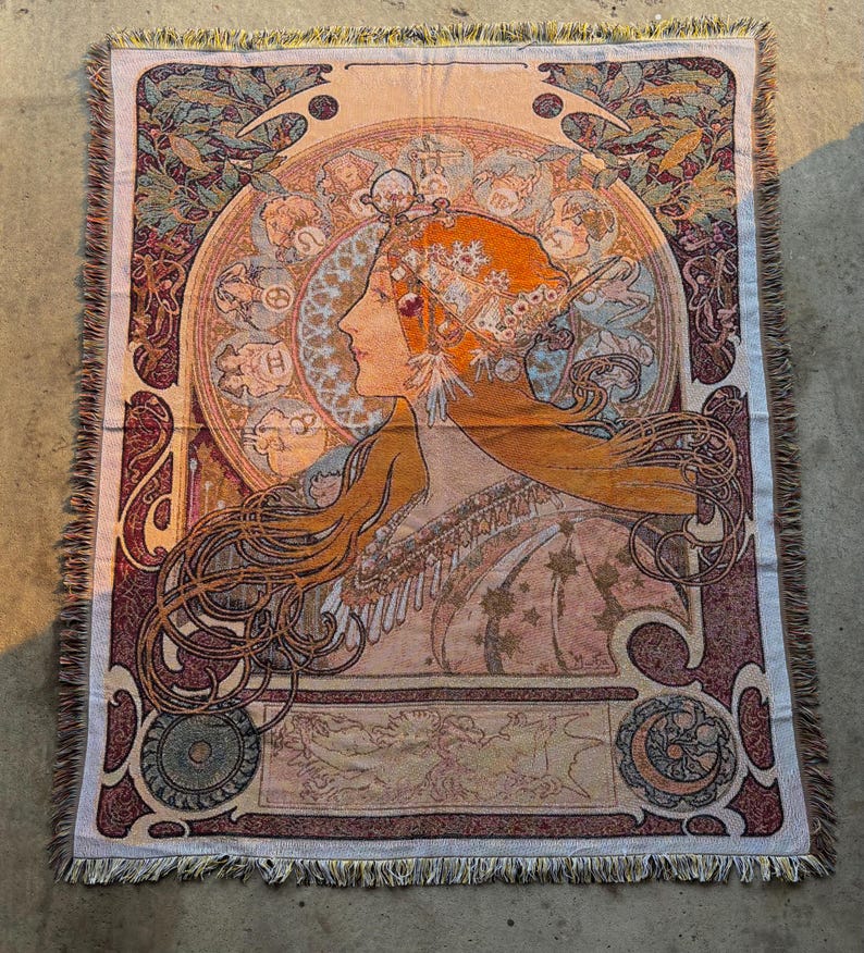 May include: Woven tapestry throw blanket featuring an Art Nouveau style design. The artwork depicts a woman's profile with flowing orange hair, surrounded by an ornate circular frame. The color palette includes shades of peach, burgundy, and teal, with a fringed edge.
