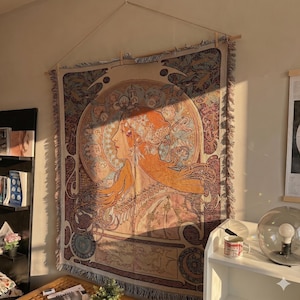 May include: A tapestry featuring a woman's profile in shades of orange, peach, and brown, with intricate Art Nouveau-style details. The artwork is displayed on a wall, adding a touch of vintage elegance to the room. The tapestry is rectangular and has a fringed edge.