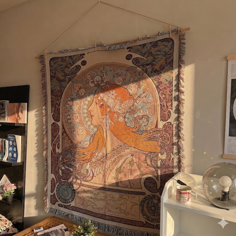 May include: A tapestry featuring a woman's profile in shades of orange, peach, and brown, with intricate Art Nouveau-style details. The artwork is displayed on a wall, adding a touch of vintage elegance to the room. The tapestry is rectangular and has a fringed edge.