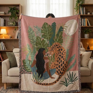 May include: A woven throw blanket with a pink background featuring a woman, a leopard, and tropical foliage. The design includes a full moon and fringed edges. The leopard has brown spots and the woman has long black hair.