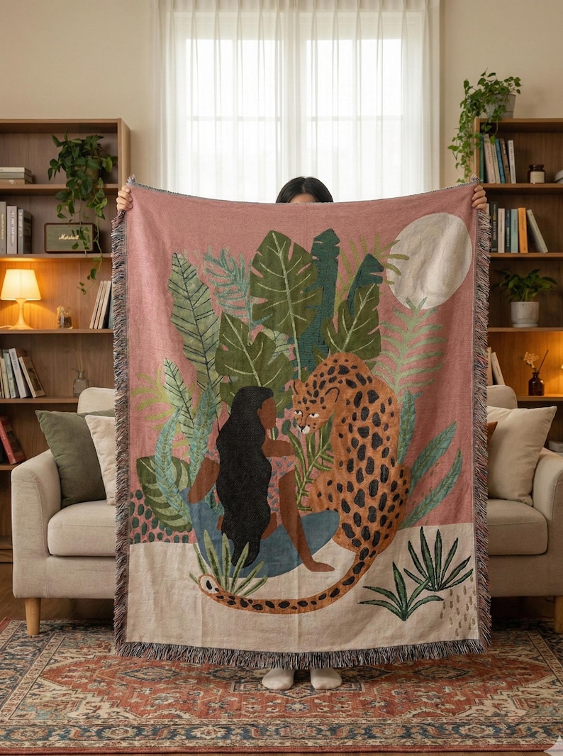May include: A woven throw blanket with a pink background featuring a woman, a leopard, and tropical foliage. The design includes a full moon and fringed edges. The leopard has brown spots and the woman has long black hair.