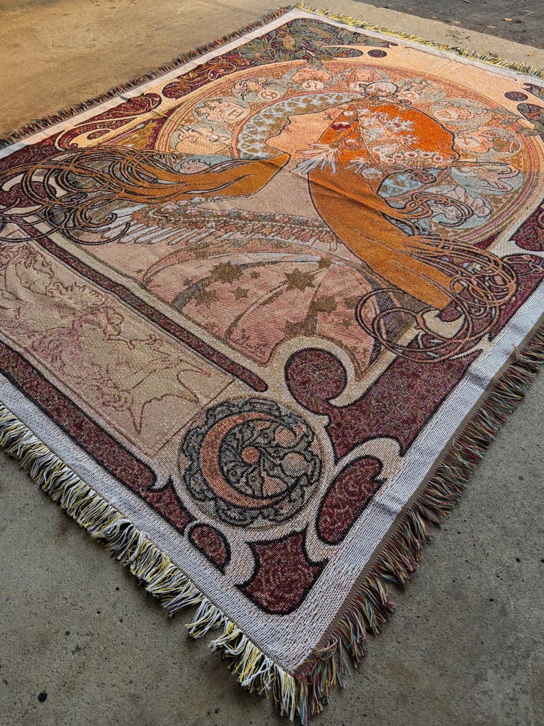 May include: Woven throw blanket featuring an Art Nouveau-style illustration of a woman with flowing red hair. The design incorporates a circular motif with stars, crescent moons, and intricate floral patterns. The color palette includes shades of orange, brown, and cream.