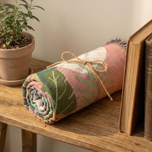 May include: Rolled-up floral throw blanket with a pink and green pattern, tied with twine. The blanket features a botanical design with leaves and flowers. The rolled blanket is resting on a wooden shelf, next to a potted plant and a book.