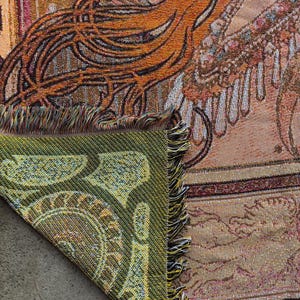 May include: Woven tapestry blanket with a detailed design in shades of orange, brown, and beige. The corner is folded over, revealing a green paisley pattern. The blanket has a fringed edge.