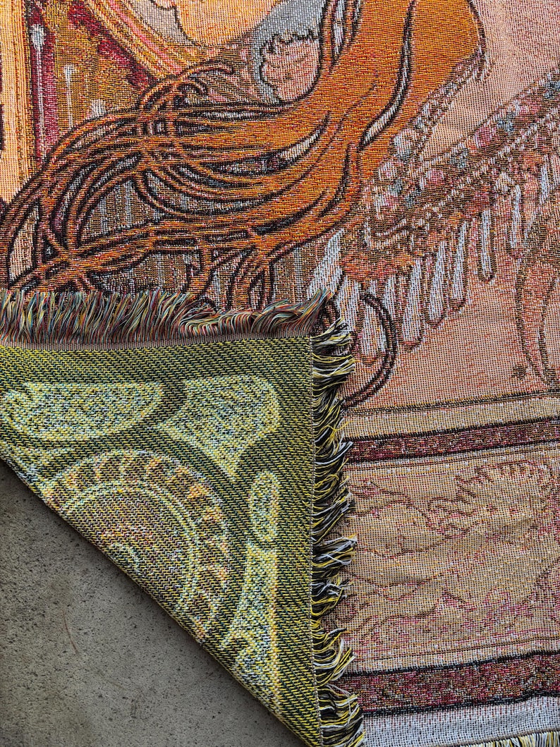 May include: Woven tapestry blanket with a detailed design in shades of orange, brown, and beige. The corner is folded over, revealing a green paisley pattern. The blanket has a fringed edge.