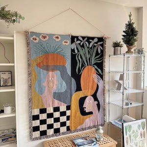 May include: A large, colourful tapestry with an abstract design. The artwork features figures, plants, and geometric shapes in shades of blue, orange, black, and yellow. The tapestry is hung on a wooden rod with a string.