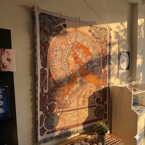 May include: A tapestry featuring a woman's profile in warm tones of orange, gold, and brown. The artwork has a circular design with floral and zodiac elements. The tapestry is rectangular with a fringed edge, hung on a wooden rod.