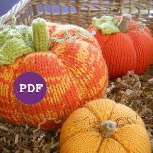 May include: Three knitted pumpkins in a basket filled with brown paper shreds. The pumpkins are orange, yellow, and a mix of orange and yellow. The largest pumpkin has a label that says "PDF".