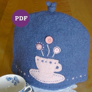 Puede incluir: Cobertor de tetera azul tejido con una taza de té rosa y flores bordadas. El cobertor tiene un detalle de botón y el texto "PDF" es visible en la esquina superior izquierda.