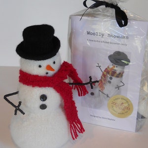May include: A handmade snowman with a black top hat, red scarf, and black bead arms. The snowman has a carrot nose and black button eyes. A package with a "Woolly Snowman" pattern is behind the snowman.