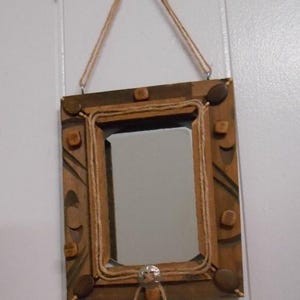 May include: A rectangular wooden mirror frame featuring a natural brown finish with carved geometric patterns. The frame is decorated with small wooden beads and a central silver-toned metal accent, all held together by a length of tan twine.