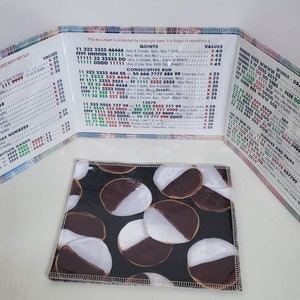 May include: A close-up of a small, folded fabric wallet with a black and white cookie pattern on the front. The wallet is open to reveal a chart with numbers and symbols, likely for a game or activity.