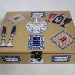 May include: A gold wooden box decorated with Passover symbols, including a Seder plate, matzah, wine glasses, and candles. The box has a hinged lid and a lock. The text "Seder Plate" and "Matzah" are visible on the box.