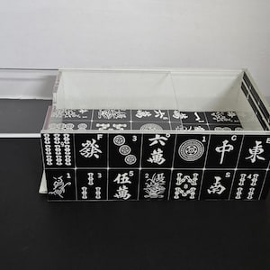 May include: A clear acrylic box with a sliding lid, filled with black and white Mahjong tiles. The box has a black exterior with white Chinese characters and symbols. The lid is partially open.