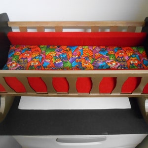 May include: A wooden doll cradle with a red and gold finish. The cradle has a colorful cat-themed mattress and a red fabric lining.