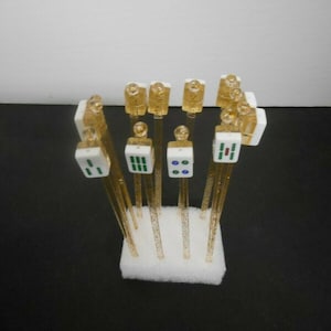 May include: A collection of decorative Mahjong tile hair sticks. The sticks are clear with gold glitter, and the tiles are white with various colored markings. The sticks are arranged in a white foam base.