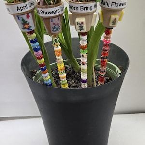May include: A black pot with green plants and four decorative flower stakes. Each stake has a small pot with artificial flowers and the text "April Showers", "Flowers Bring", and "Mahj Flowers". The stakes are adorned with colorful beads.