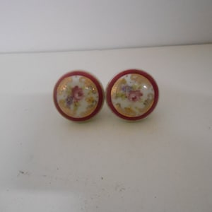 May include: Two round ceramic knobs with a red rim and a white center. The center features a floral design with gold accents.