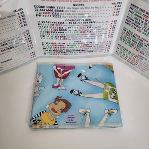 May include: A blue fabric wallet with a white border and a colorful cartoon design of women playing golf. The wallet has a small white tag that says "Created especially for you by King of Albany".